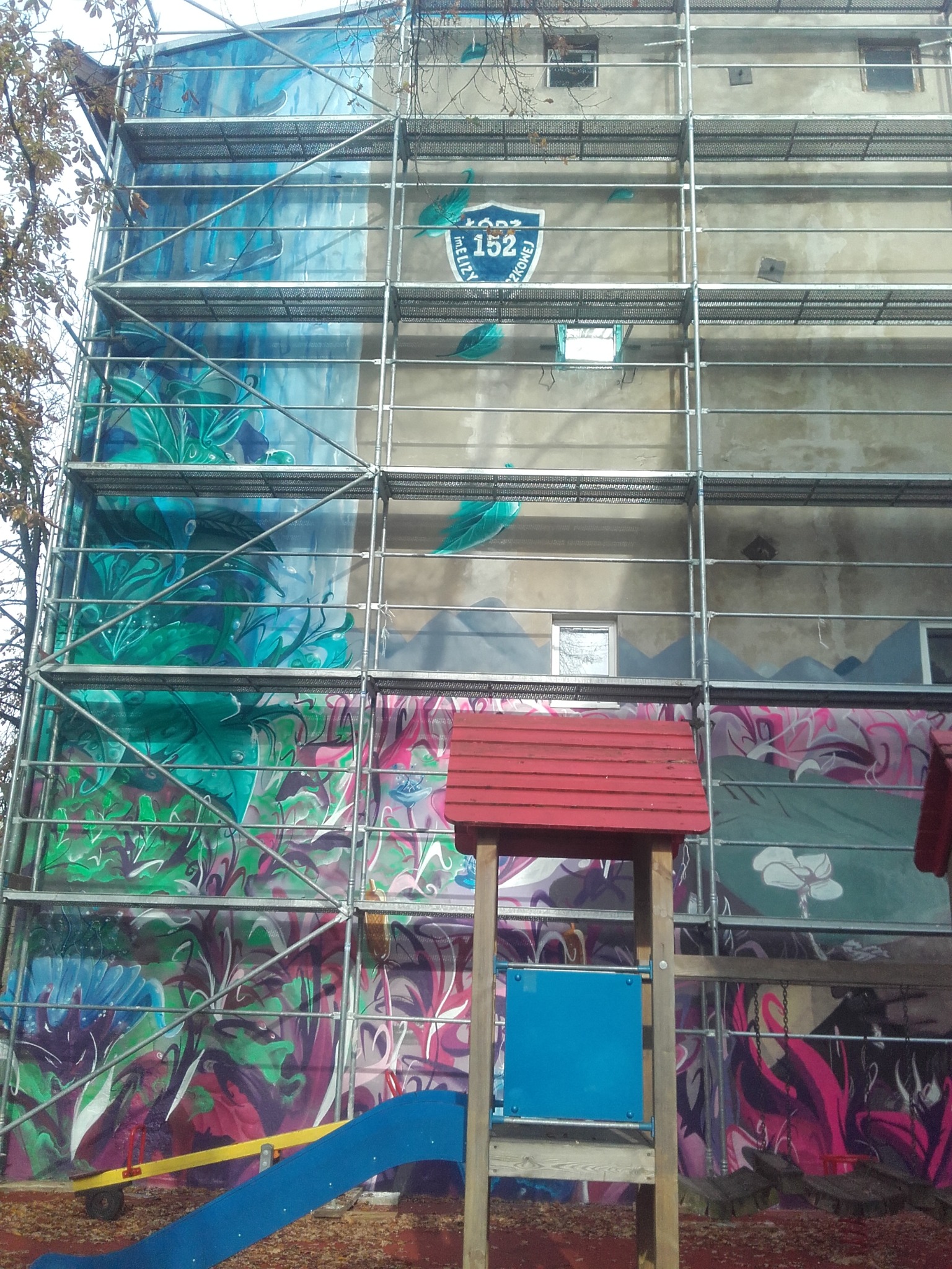 Budynek w rusztowaniach z muralem w trakcie tworzenia, widoczny fragment placu zabaw. Ujęcie z dołu, podkreślające wysokość i etap prac.