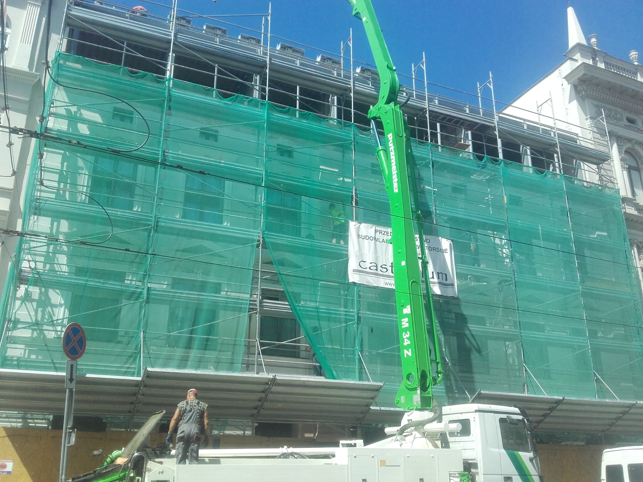 Elewacja budynku w rusztowaniach, osłonięta zieloną siatką. Na pierwszym planie ramię pompy do betonu Putzmeister M 34 Z i pracownik budowlany na platformie.