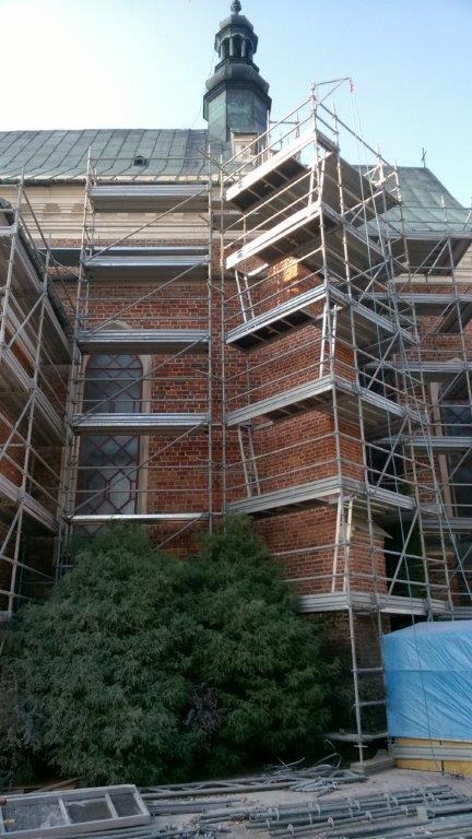 Renowacja ceglanej elewacji kościoła z wieżą, otoczonego rusztowaniami. Widoczne okna z witrażami i zieleń. Prace elewacyjne w toku, z dbałością o detale architektoniczne.