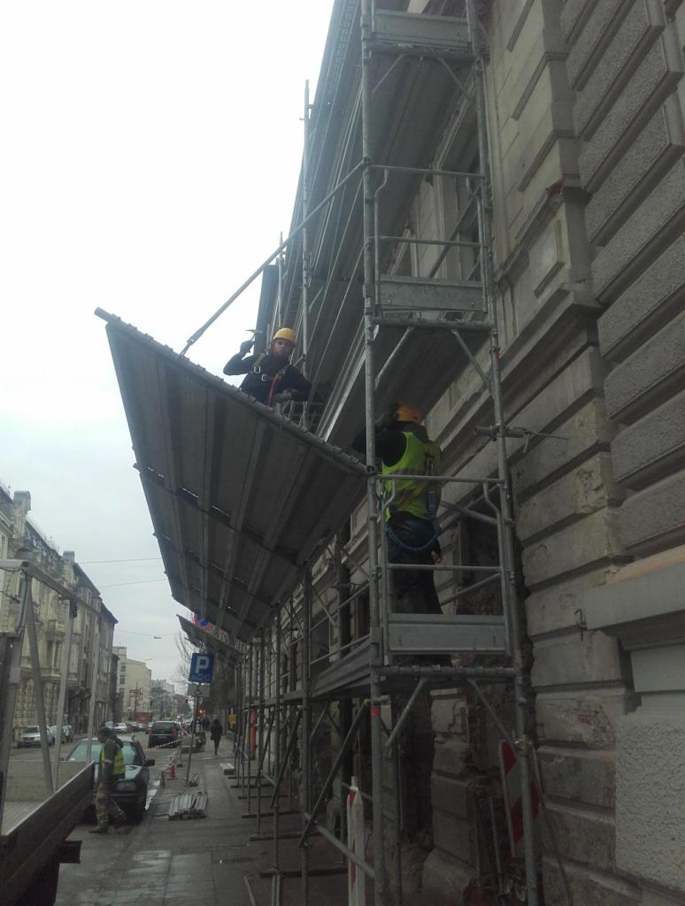 Pracownicy na rusztowaniu przy budynku z elewacją w trakcie renowacji; widoczne detale architektoniczne i zabezpieczenia.