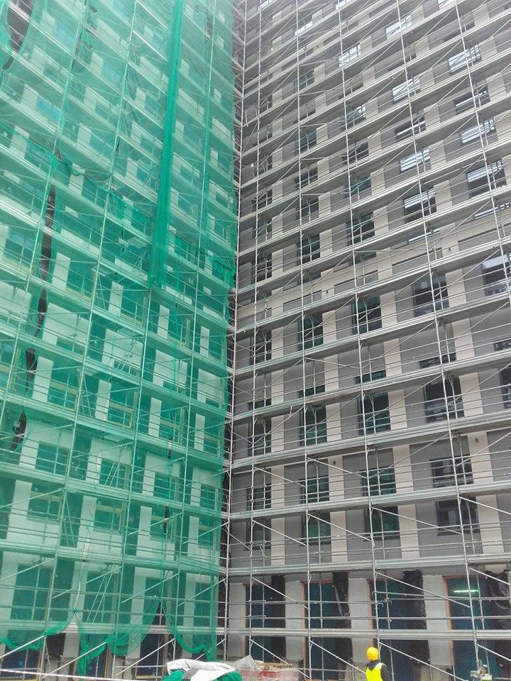 Budynek w trakcie prac elewacyjnych, widoczne rusztowania z siatką ochronną po lewej stronie i elewacja po prawej, pracownik w żółtym kasku na dole.