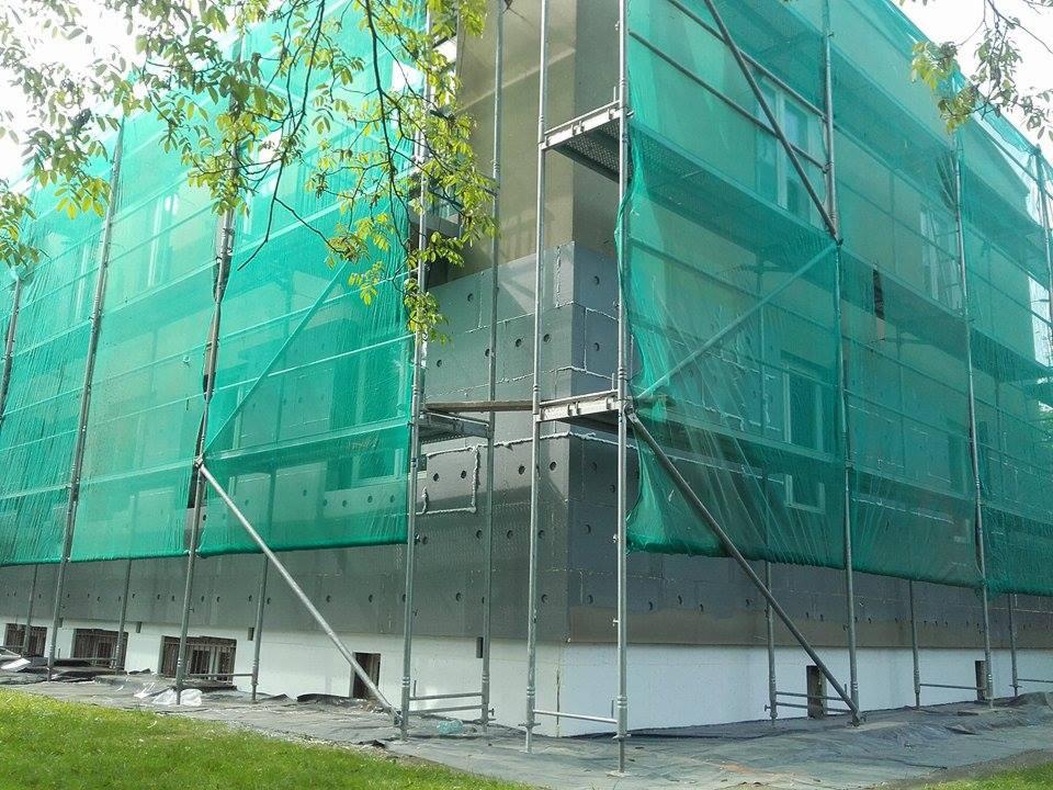 Budynek w trakcie ocieplania, zabezpieczony rusztowaniem i siatką ochronną, widoczne szare płyty izolacyjne z otworami montażowymi na elewacji.
