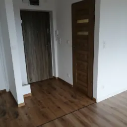 Nowoczesne wnętrze przedpokoju z dwiema drewnianymi drzwiami: wejściowymi i wewnętrznymi, oraz świeżo ułożonymi panelami podłogowymi w odcieniach brązu, widoczne listwy przypodłogowe.