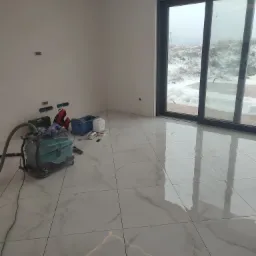 Wnętrze w trakcie prac wykończeniowych: białe płytki podłogowe z marmurkowym wzorem, odkurzacz przemysłowy, narzędzia i akcesoria budowlane, duże przesuwne okno z widokiem na zasypany śniegiem...