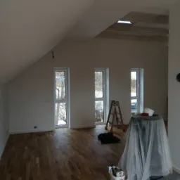 Wnętrze poddasza po malowaniu ścian na biało, z drewnianą podłogą, trzema oknami, drewnianą drabiną malarską i wiadrem z farbami w rogu.