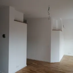 Puste, świeżo wyremontowane wnętrze z drewnianą podłogą, białymi ścianami z wystającymi elementami konstrukcyjnymi, widocznymi gniazdkami elektrycznymi i niedokończonym oświetleniem sufitowym.