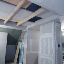 Montaż płyt kartonowo-gipsowych na suficie i ścianach, widoczne drewniane belki konstrukcyjne i otwory w suficie, pomieszczenie w trakcie prac wykończeniowych, szpachlowane łączenia płyt.