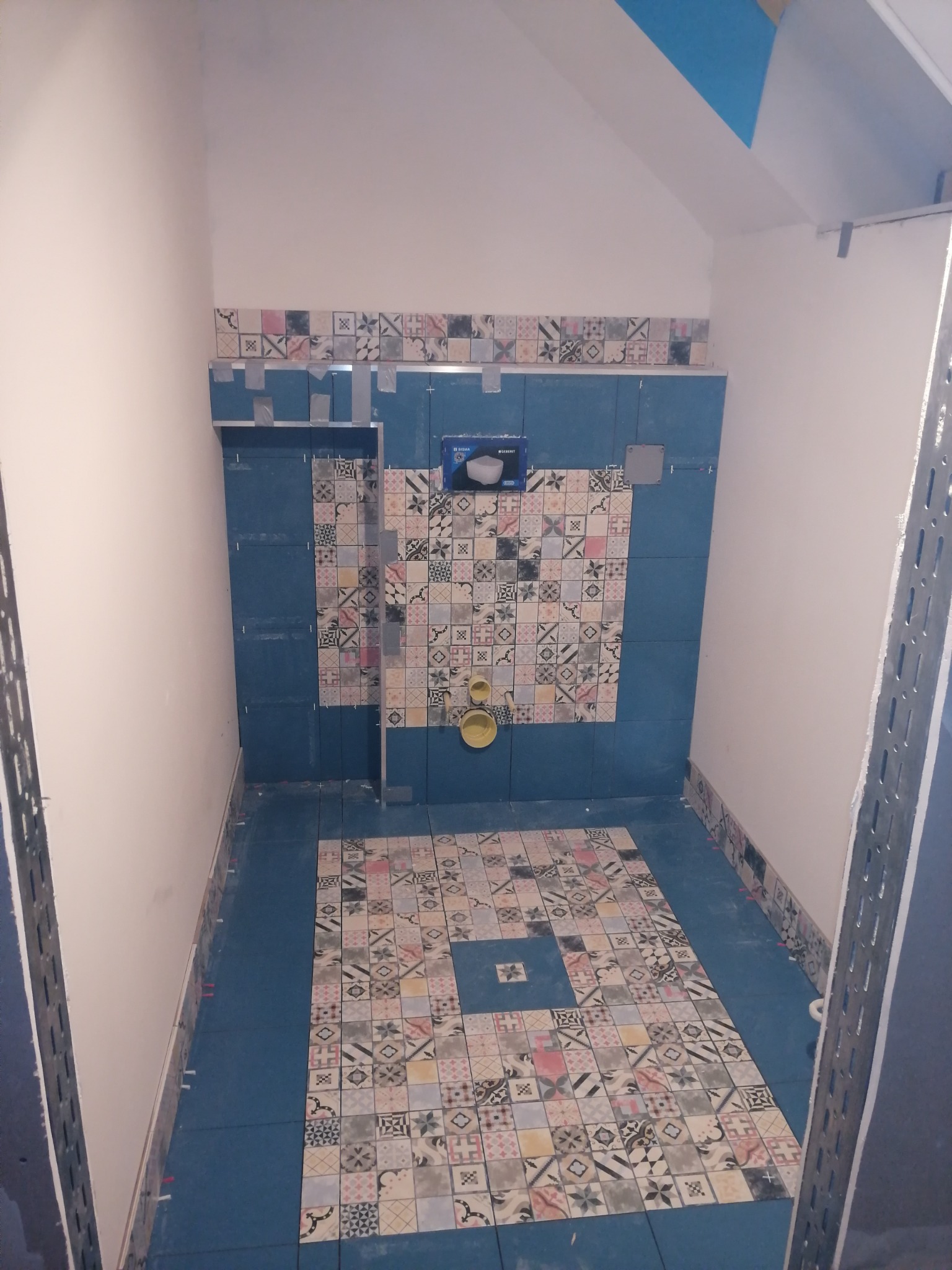 Niedokończona łazienka z niebieskimi płytkami i wzorzystą mozaiką, widoczne elementy instalacji i metalowe profile montażowe.