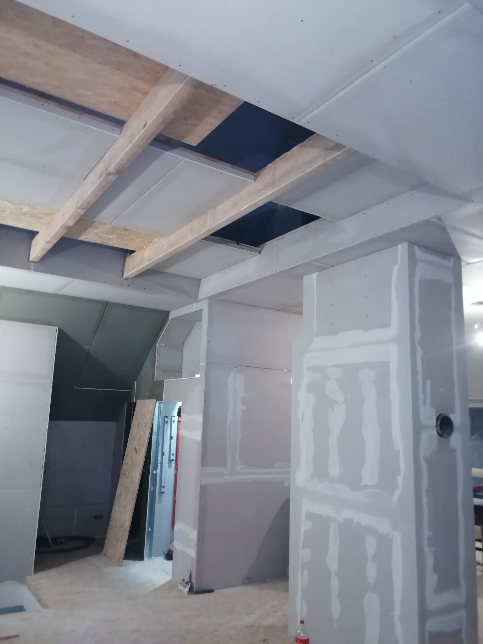 Montaż płyt kartonowo-gipsowych na suficie i ścianach, widoczne drewniane belki konstrukcyjne i otwory w suficie, pomieszczenie w trakcie prac wykończeniowych, szpachlowane łączenia płyt.