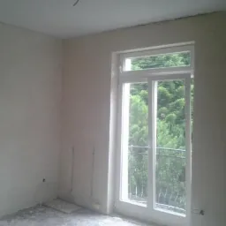 Pokój w trakcie remontu, ściany po nałożeniu gładzi, surowa podłoga, okno balkonowe z widokiem na zieleń.