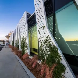 Nowoczesny budynek usługowy z ażurową, białą elewacją o geometrycznym wzorze trójkątów, porośniętą pnącą roślinnością i ozdobną trawą, widziany z perspektywy chodnika.