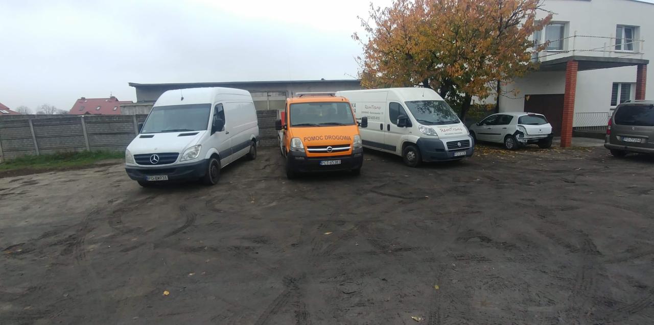 Trzy busy, w tym jeden pomarańczowy z napisem 'Pomoc Drogowa', stoją na błotnistym parkingu przed budynkiem.