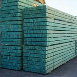 Stos impregnowanych na zielono desek drewnianych ułożonych w równe warstwy, widoczny fragment nieba w tle.
