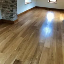 Jasna, lakierowana podłoga drewniana z wyraźnym usłojeniem, ułożona wzdłuż pomieszczenia, z fragmentem kamiennej kolumny po lewej stronie i widocznymi oknami w tle.