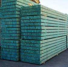 Stos impregnowanych na zielono desek drewnianych ułożonych w równe warstwy, widoczny fragment nieba w tle.