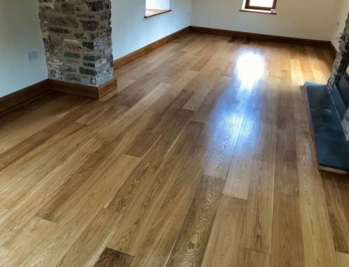 Jasna, lakierowana podłoga drewniana z wyraźnym usłojeniem, ułożona wzdłuż pomieszczenia, z fragmentem kamiennej kolumny po lewej stronie i widocznymi oknami w tle.