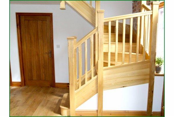 Jasne, drewniane schody z balustradą w nowoczesnym wnętrzu domu, widoczne drzwi z tego samego drewna i podłoga, niewielka roślina doniczkowa przy oknie.