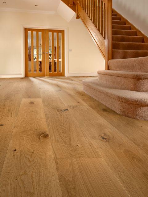 Jasna, drewniana podłoga z wyraźnym usłojeniem, prowadząca w stronę dwuskrzydłowych, przeszklonych drzwi i schodów obłożonych beżową wykładziną.