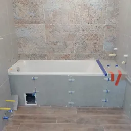 Łazienka w trakcie wykańczania: obudowa wanny z szarych płytek, płytki wzorzyste na ścianie, drewnopodobna podłoga, widoczne krzyżyki dystansowe i taśma malarska.