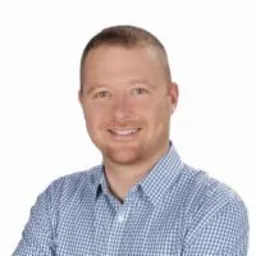 Uśmiechnięty mężczyzna w niebieskiej koszuli w kratę na jasnym tle. Portret biznesowy z delikatnym oświetleniem, ukazujący profesjonalny wizerunek.