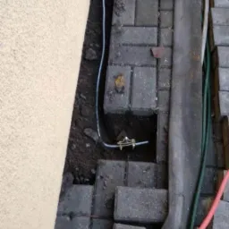 Montaż instalacji elektrycznej przy budynku: widoczne przewody i złącze w wykopie obok kostki brukowej.