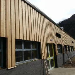 Budynek z elewacją z pionowych desek drewnianych, szare okna i ciemna podmurówka, na tle wzgórza i pochmurnego nieba.