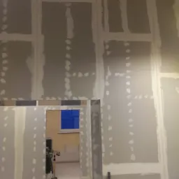 Wnętrze w trakcie montażu płyt gipsowo-kartonowych na ścianach, widoczne szpachlowane łączenia, metalowa drabina i lampa budowlana na trójnogu, w tle okno z niebieskimi roletami.