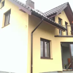 Żółta elewacja dwukondygnacyjnego domu z brązowymi oknami, dachem i rynnami, balkon z kutą balustradą, widoczne materiały budowlane przy wejściu.