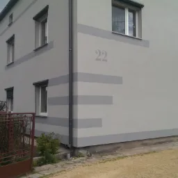 Szary dom jednorodzinny z numerem 22 na elewacji i poziomymi pasami, widok z zewnątrz, brama w tle.
