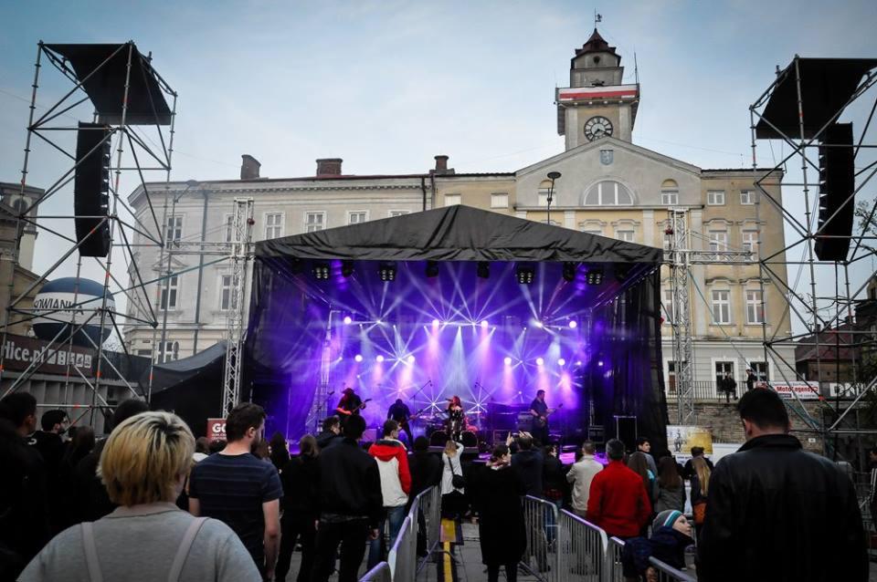 Scena plenerowa z zespołem muzycznym oświetlona fioletowymi światłami, widoczna publiczność i rusztowania nagłośnieniowe na tle budynku z wieżą zegarową.