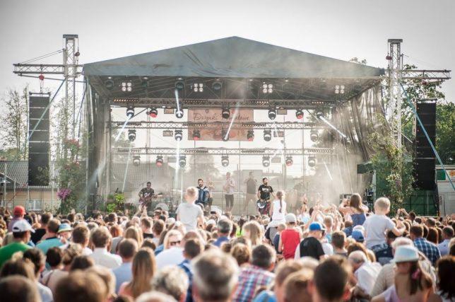 Scena plenerowa z zespołem muzycznym podczas koncertu dla dużej publiczności, z widocznym nagłośnieniem i oświetleniem.