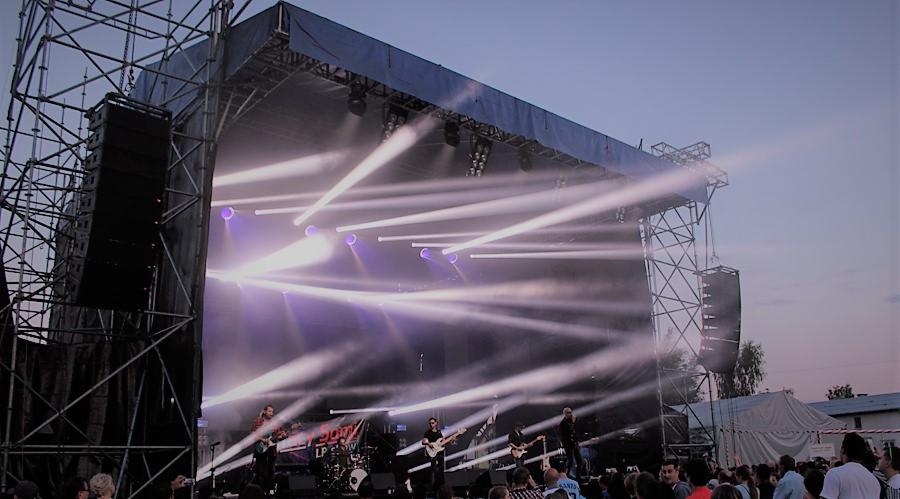 Scena koncertowa z zespołem muzycznym oświetlona reflektorami i nagłośniona, widoczna publiczność.