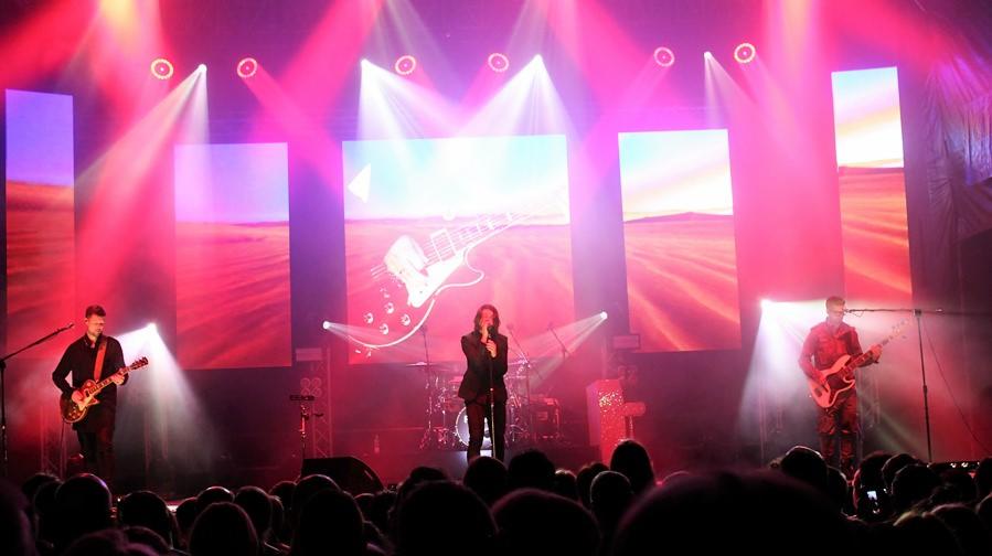 Zespół muzyczny na scenie podczas koncertu, oświetlony reflektorami, z publicznością w tle.