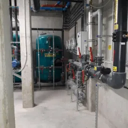 Kompleksowa instalacja wodna z filtrami piaskowymi, rurami PCV z czerwonymi zaworami, oraz elementami sterującymi w pomieszczeniu technicznym.