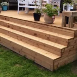 Drewniane schody tarasowe z czterema stopniami prowadzące na taras z białymi meblami ogrodowymi i roślinami w donicach.