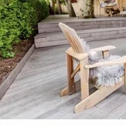 Drewniany taras z krzesłem Adirondack wyłożonym szarą skórą, schody prowadzące do podwyższonej części tarasu z widocznym pniem drzewa w tle.