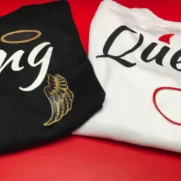 Dwa swetry leżące obok siebie na czerwonym tle: czarny z białym napisem 'King' ozdobionym złotym aureolą i skrzydłami, oraz biały z czarnym napisem 'Queen' i czerwonymi rogami oraz ogonem.
