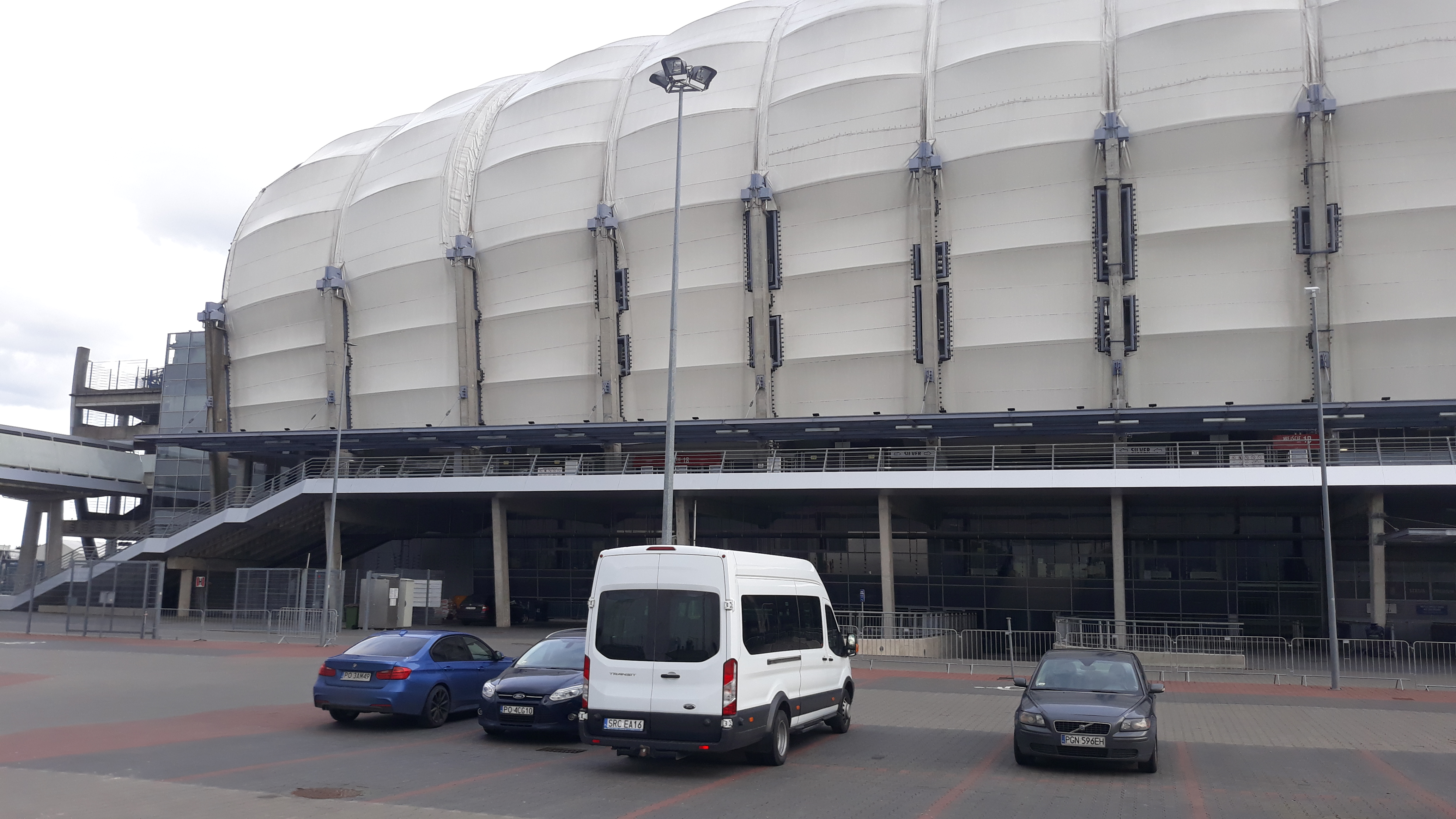 Biały bus transportowy zaparkowany przed stadionem, obok zaparkowane samochody osobowe.