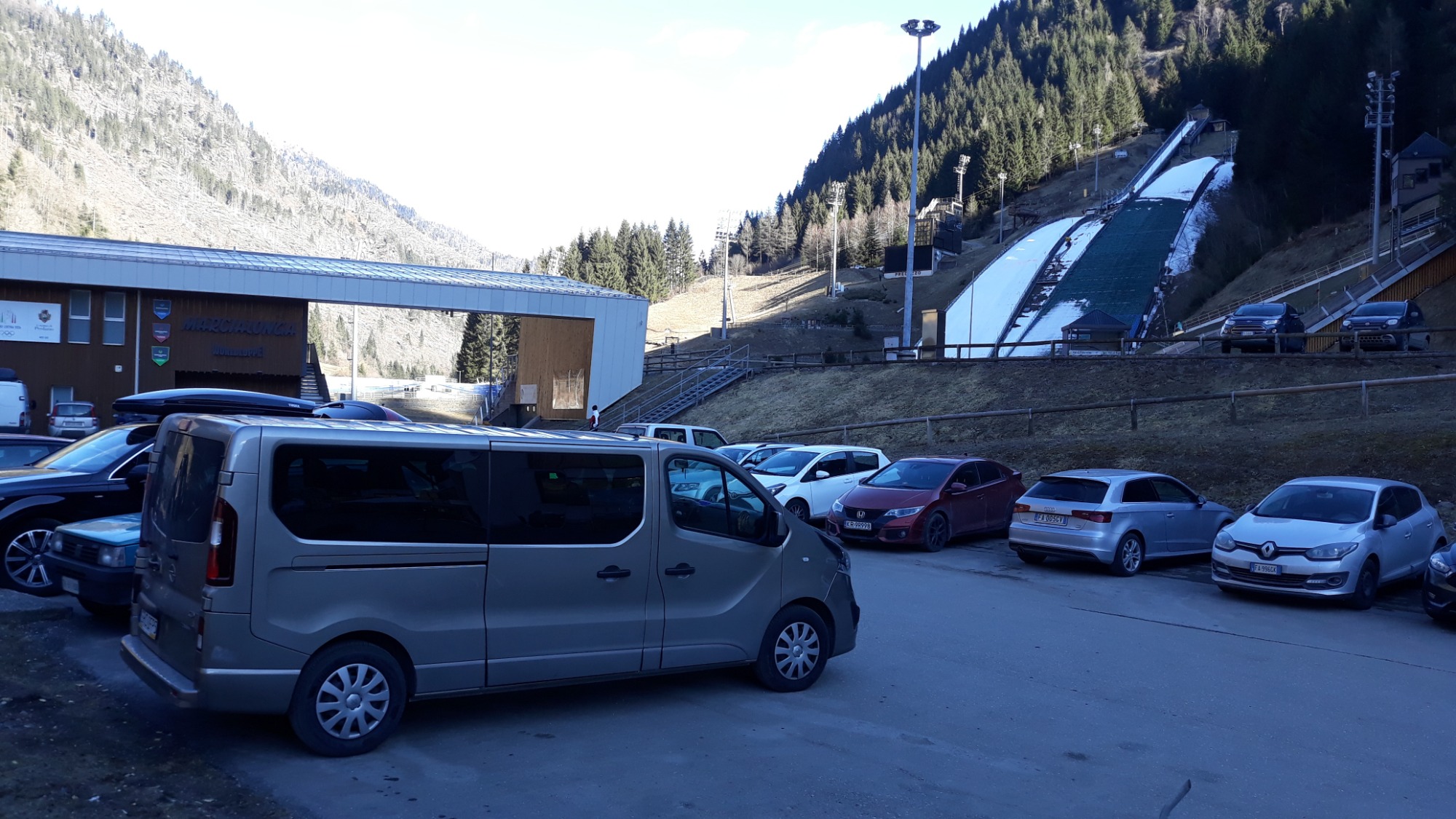 Szary van na parkingu przed skocznią narciarską w słoneczny dzień; w tle góry i las