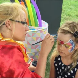 Kobieta w kostiumie maluje farbami twarz dziewczynki w plenerze, w tle kolorowe balony i wiaderko w geometryczne wzory.