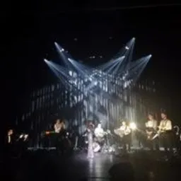 Zespół muzyczny na scenie podczas występu, oświetlony reflektorami tworzącymi rozgwieżdżony efekt, centralnie umieszczona wokalistka w jasnej sukni, pozostali muzycy w ciemnych strojach.