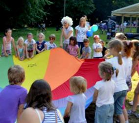 Grupa dzieci bawi się kolorową chustą animacyjną na trawie w parku, animator w białej peruce obserwuje zabawę.