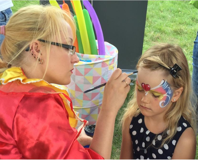 Kobieta w kostiumie maluje farbami twarz dziewczynki w plenerze, w tle kolorowe balony i wiaderko w geometryczne wzory.