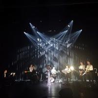 Zespół muzyczny na scenie podczas występu, oświetlony reflektorami tworzącymi rozgwieżdżony efekt, centralnie umieszczona wokalistka w jasnej sukni, pozostali muzycy w ciemnych strojach.