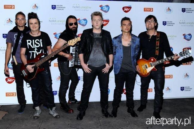 Zespół muzyczny, sześciu mężczyzn pozuje z instrumentami na tle ściany z logotypami sponsorów, gitara basowa z przodu, gitara elektryczna z prawej strony, jeden muzyk w okularach przeciwsłonecznych.