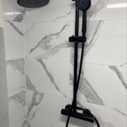 Czarny zestaw natryskowy z deszczownicą i słuchawką na tle białych płytek imitujących marmur w łazience.