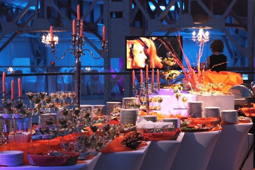 Bogato zastawiony stół z przekąskami, deserami i napojami na eleganckim przyjęciu, z dekoracjami w odcieniach pomarańczy i świecznikami, w tle konstrukcja stalowa i ekran wyświetlający wizerunek...