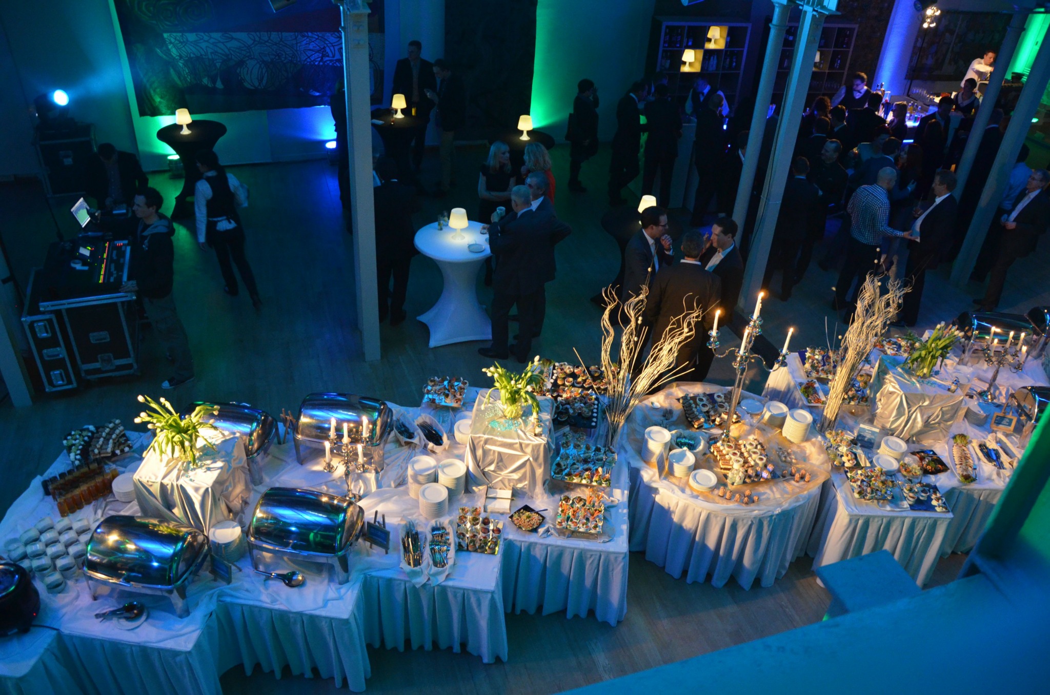 Widok z góry na stoły cateringowe z dekoracjami i potrawami podczas imprezy, w tle goście i oświetlenie w odcieniach niebieskiego i zielonego.