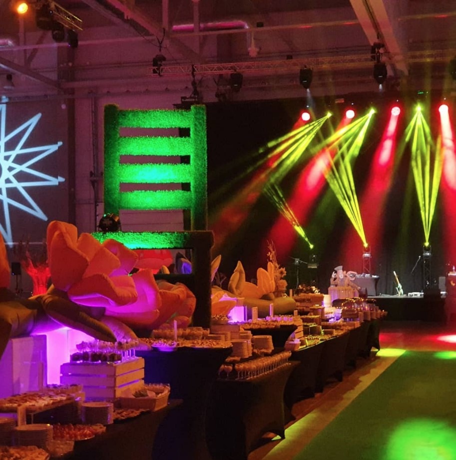 Uroczysta sala bankietowa z długim stołem cateringowym, ozdobiona kwiatami i dekoracjami z trawy, oświetlona reflektorami scenicznymi.