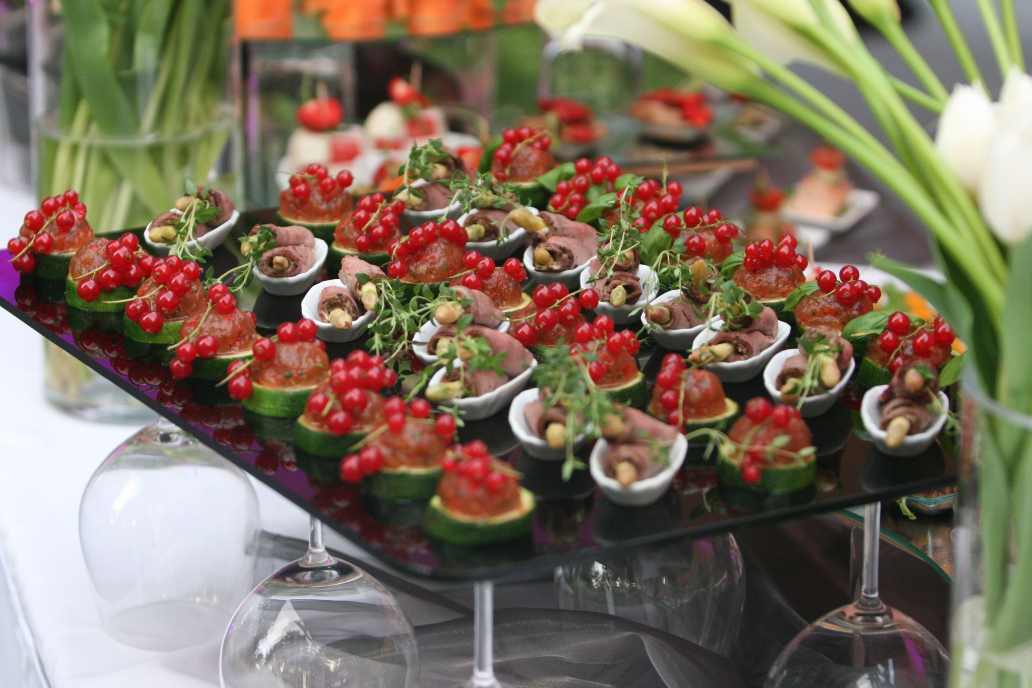 Elegancko podane przekąski na czarnym, lustrzanym podeście: plasterki cukinii z czerwoną galaretką i porzeczkami oraz roladki mięsne w białych, małych miseczkach, dekorowane zielonymi ziołami,...